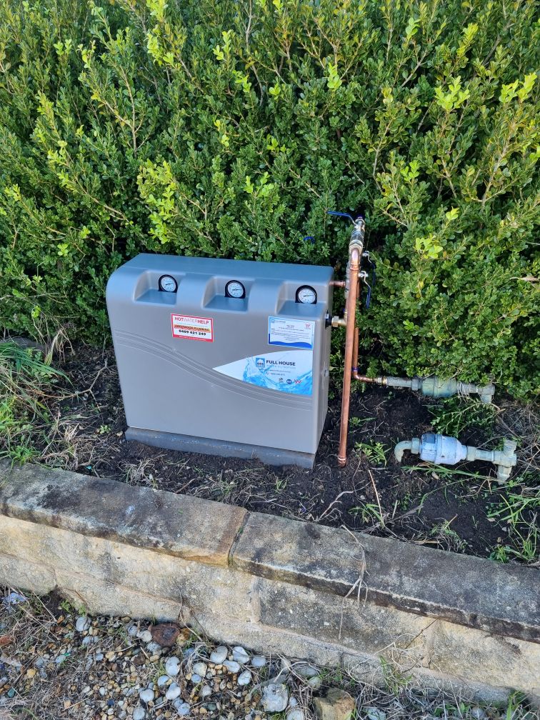 Grey water pump system with pipes installed on soil beside a hedge and a stone border.