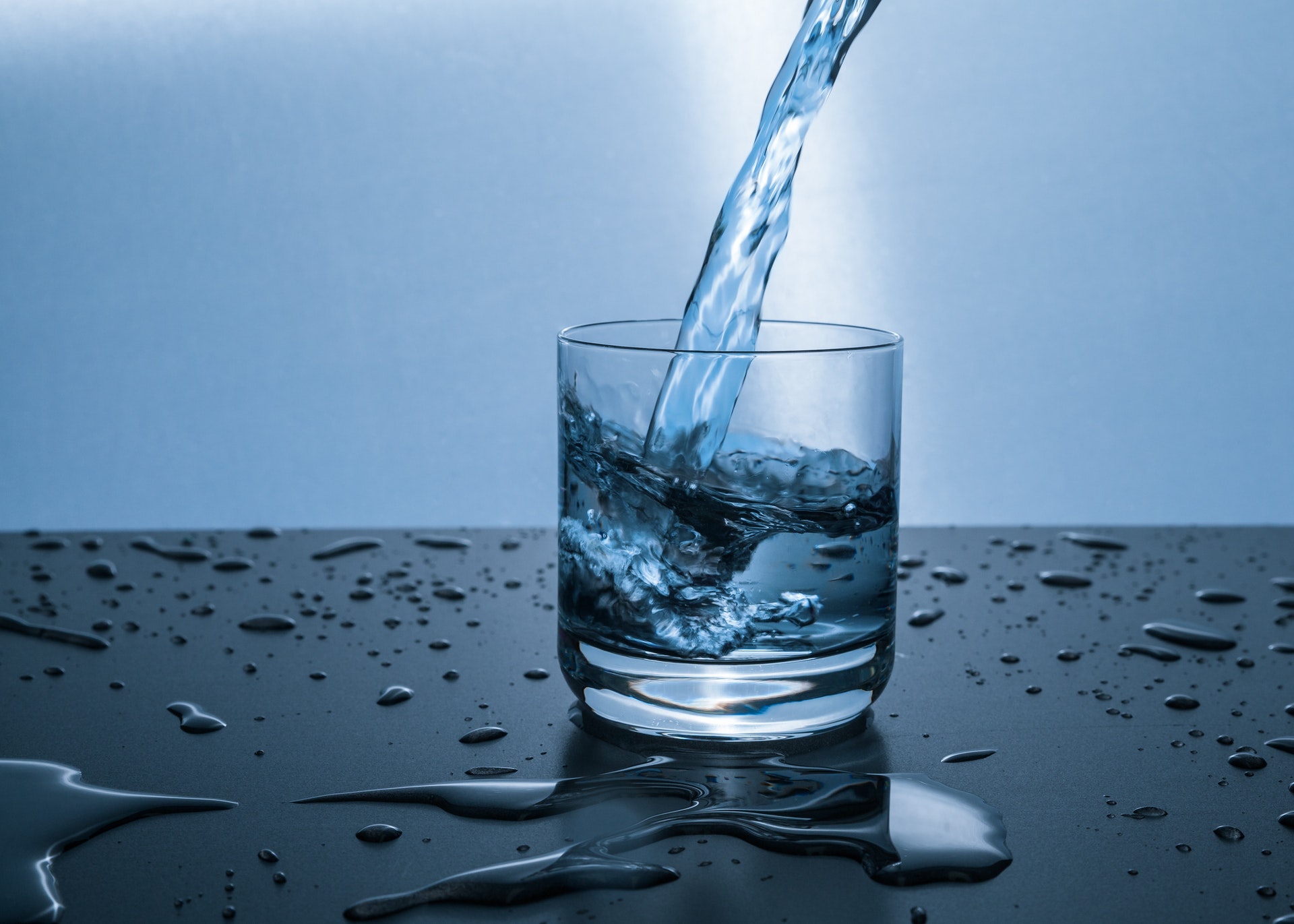 Water being poured into a clear glass, surrounded by water droplets on a dark surface with a blue-toned background.