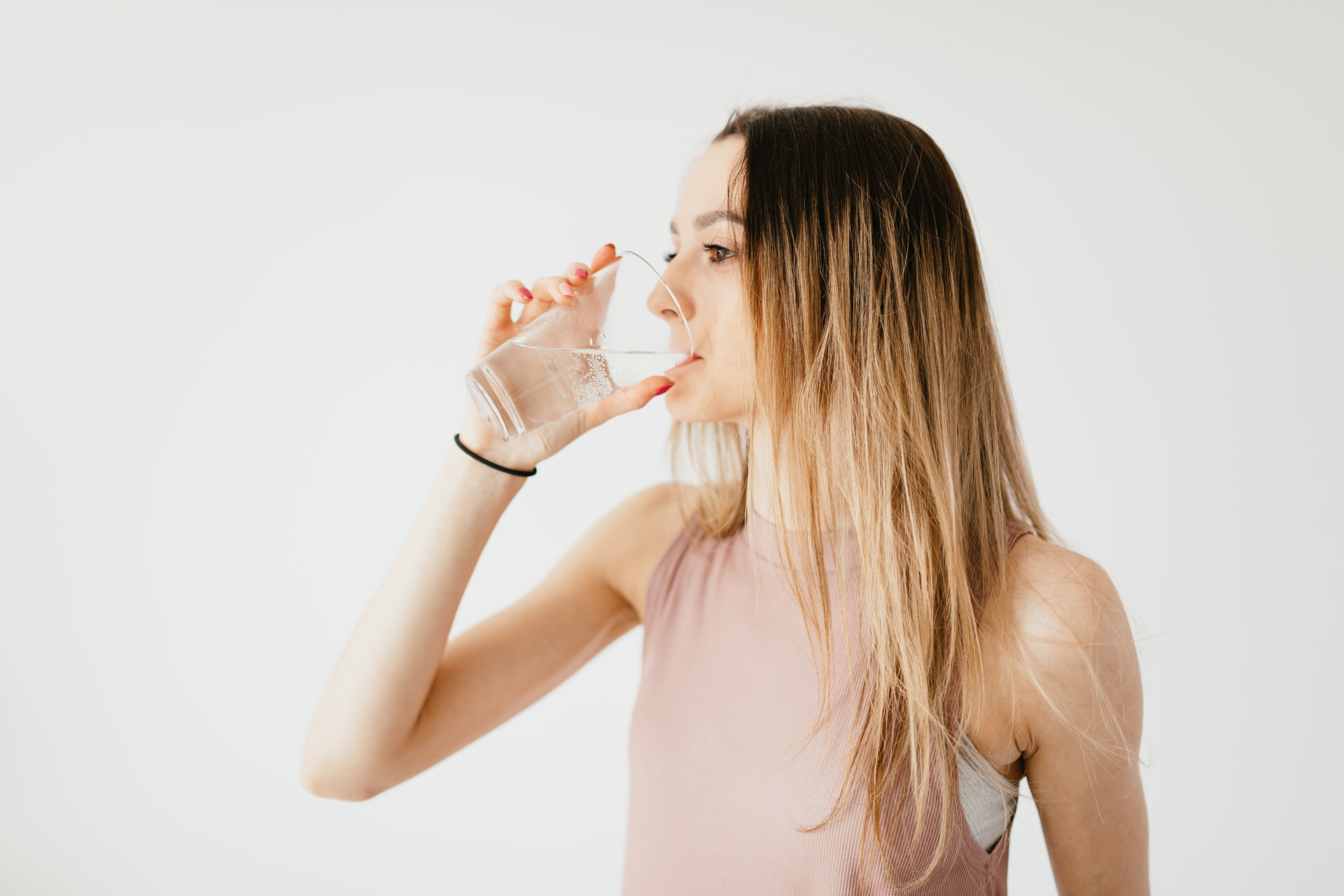 Woman with long hair wearing a sleeveless top drinks alkaline water from a clear glass against a plain white background.
