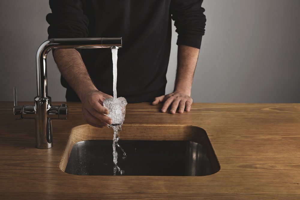 A person holds a glass under a running kitchen faucet with an Alkaline Water Filter, filling it with water over a wooden countertop and sink for performance & recovery support.