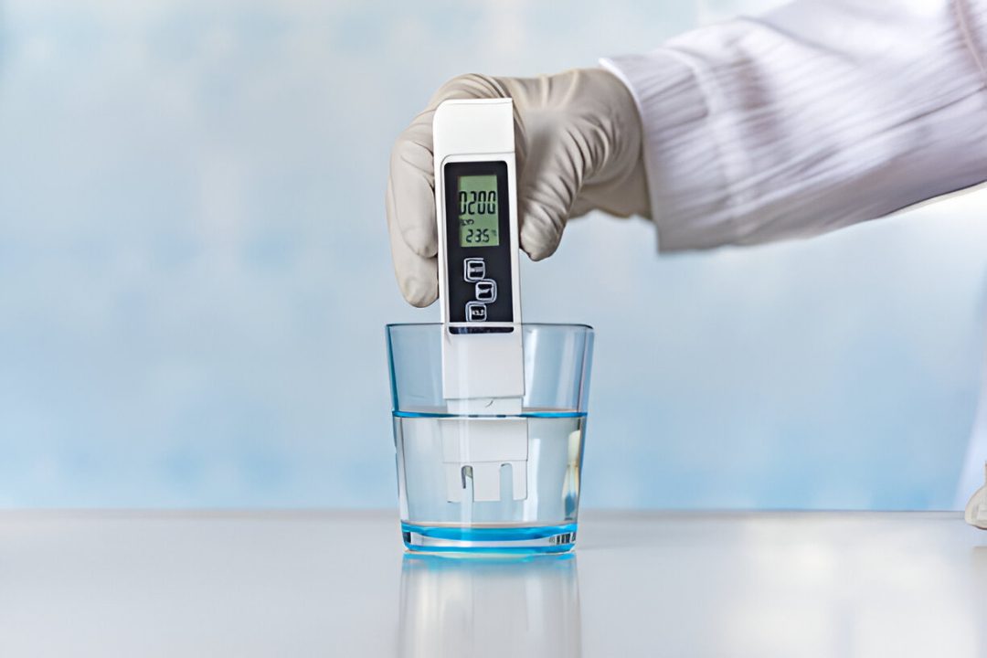Gloved hand holding a digital water quality tester in a glass of water, with TDS tested measurements shown on the screen.