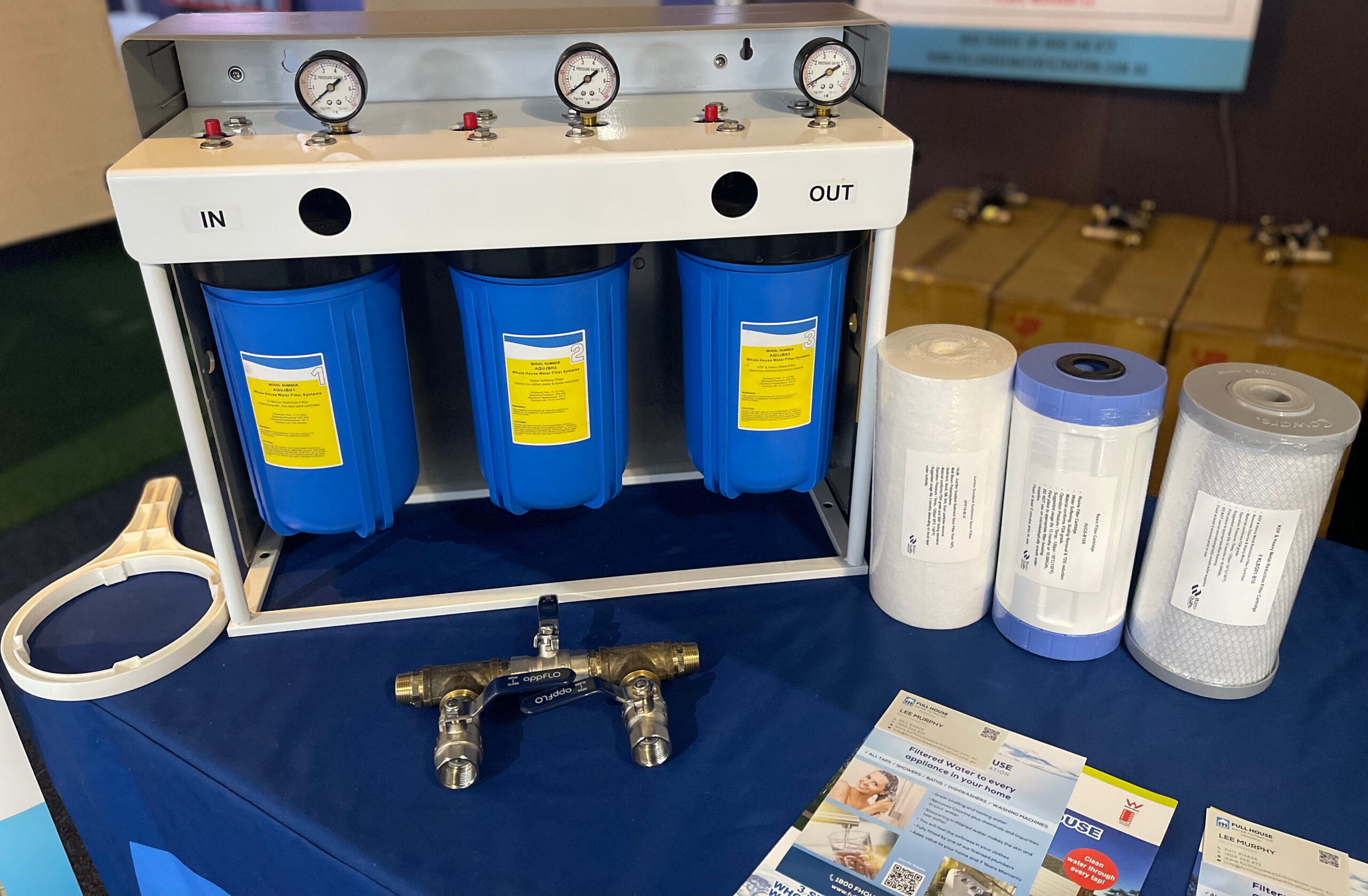 Three blue water filter cartridges in a white frame, labeled IN and OUT, with top gauges and red control buttons.